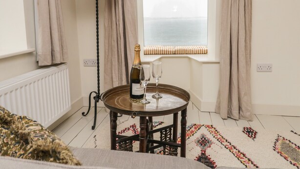 A room with a round wooden table, a bottle of champagne, and two glasses. There is a window with curtains, a radiator, and a patterned rug on the floor. - Waterside (Ravenglass)