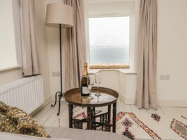 Um quarto com uma mesa de madeira redonda, uma garrafa de champanhe e duas taças. Há uma janela com cortinas, um radiador e um tapete estampado no chão.