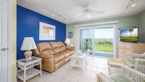 A living room with a brown leather sofa, white wicker furniture, and a television on a wooden cabinet. - Four Winds by Distinctive Beach Rentals (St. Augustine)