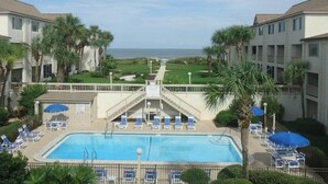A pool area with lounge chairs and umbrellas, surrounded by buildings with a view of the beach. - Four Winds by Distinctive Beach Rentals (St. Augustine)