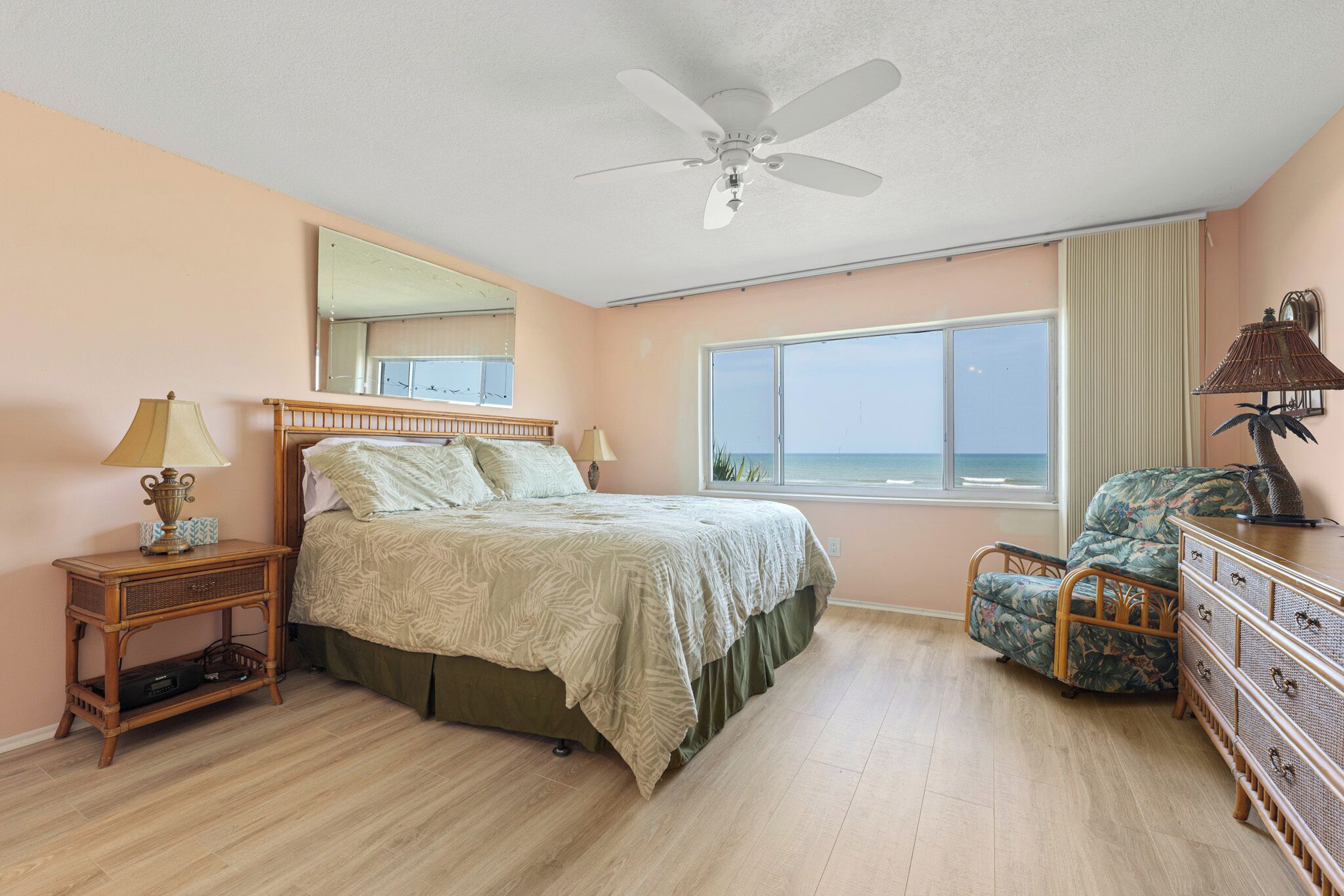 Ein Schlafzimmer mit Bett, Nachttisch, Sessel, Kommode, Deckenventilator und einem großen Fenster mit Meerblick.