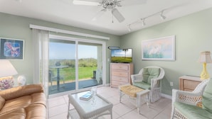A spacious living area with a sliding glass door leading to a balcony, wicker furniture, a ceiling fan, and artwork on the walls. - Four Winds by Distinctive Beach Rentals (St. Augustine)