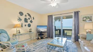 A living room with a TV, a glass coffee table, a sofa, a chair, and a balcony with a view of the ocean.