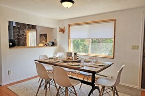 A dining area with a wooden table set for four, white chairs, a stone wall, a window with blinds, and a ceiling light. - Great Backyard with Firepit, Wood Burning Stove & Pet Friendly! (Grand Lake)