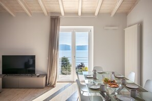 A modern living room with a flat-screen TV, a glass dining table set for two, and a large sliding glass door leading to a balcony with a view of the sea.