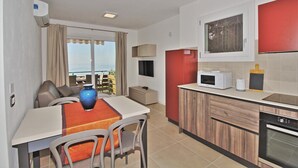 A compact kitchen with a microwave, a coffee maker, and a toaster. There is a dining table with chairs and a vase. The kitchen has a red accent wall and a view of the outdoors.