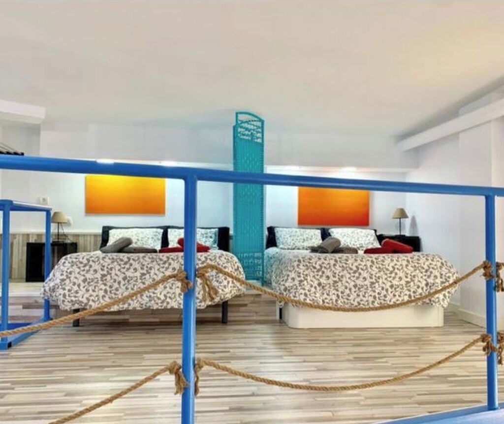 Quarto de hotel com duas camas, piso de madeira, corrimão azul e barreira de corda.