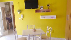 Un comedor con una mesa blanca y cuatro sillas, un televisor de pantalla plana montado en una pared amarilla y un estante con objetos decorativos.