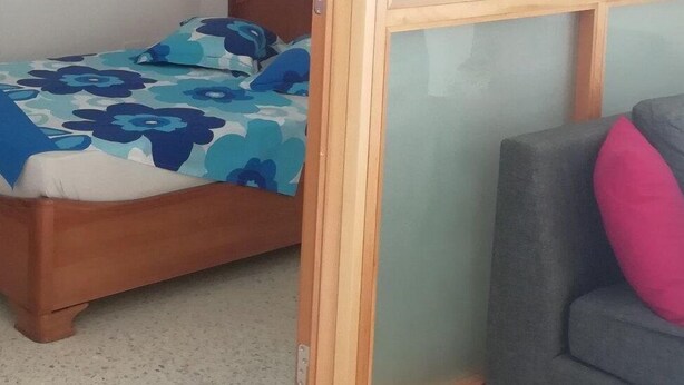 Una cama con cubrecama floral, un sofá gris con un cojín rosa y una ventana con marco de madera.