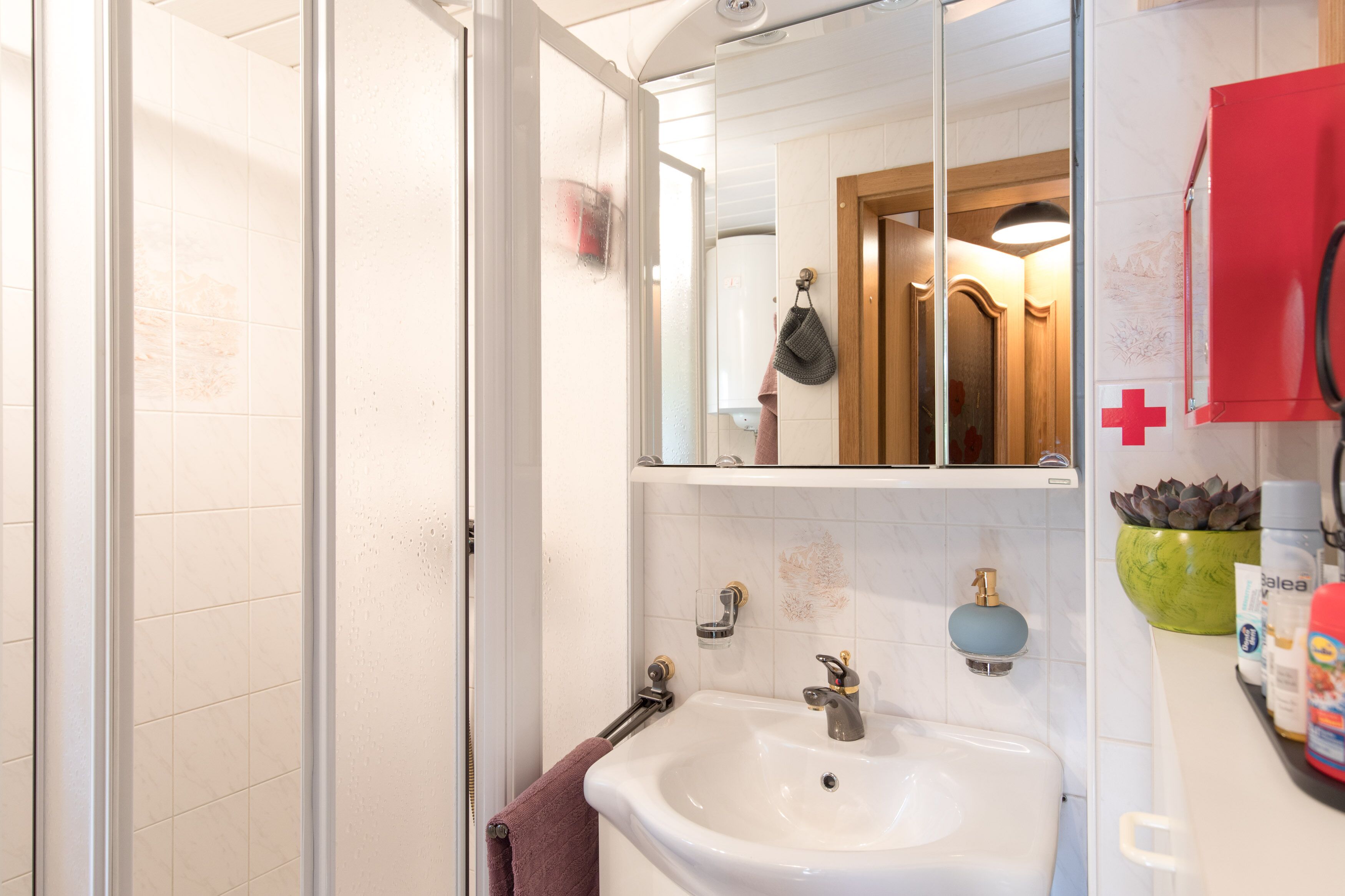 Een badkamer met een wit wastafel, een spiegel, een handdoekhouder, een douchedeur en een rode kast.