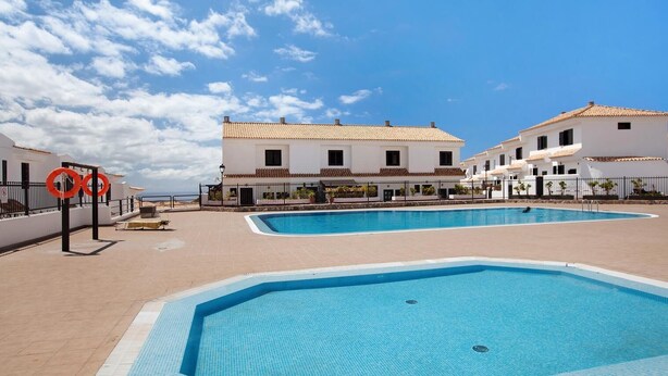 Un área de piscina con dos piscinas, rodeada de edificios blancos con techos de tejas, y un cielo azul despejado.
