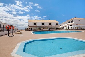 Ett poolområde med två pooler, omgivet av vita byggnader med tak belagda med tegelpannor, och en klarblå himmel.
