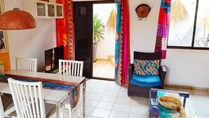 Un comedor con una mesa y sillas blancas, un televisor y un sofá de mimbre con cojines de colores.