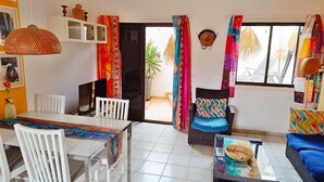 Un comedor con una mesa y sillas, una silla de mimbre con cojín, un sofá azul y una ventana con cortinas.