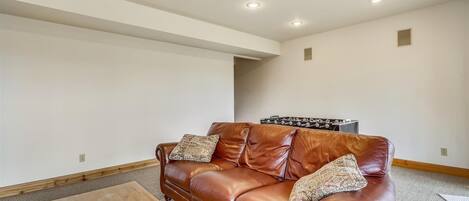 Un salón amplio con un sofá de cuero marrón, dos cojines decorativos estampados, una mesa de centro de madera y un control remoto encima.