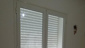 Een kamer met een groot raam, afgedekt met jaloezieën, een klein tafeltje met een lichtgekleurd tafelkleed en een vloerkleed met dessin.