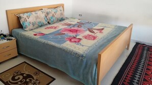 Een slaapkamer met een houten bed, een bloemenprint deken, een nachtkastje met een lamp en een kamerkleed met dessin.