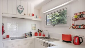 Eine Küche mit weißen Schränken, einer Edelstahl-Spüle, einem roten Wasserkocher und einem Fenster mit Blick auf Grünflächen.