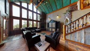 Amplia sala de estar con un gran ventanal, piso de madera, chimenea y una pared de piedra con una escalera de madera.