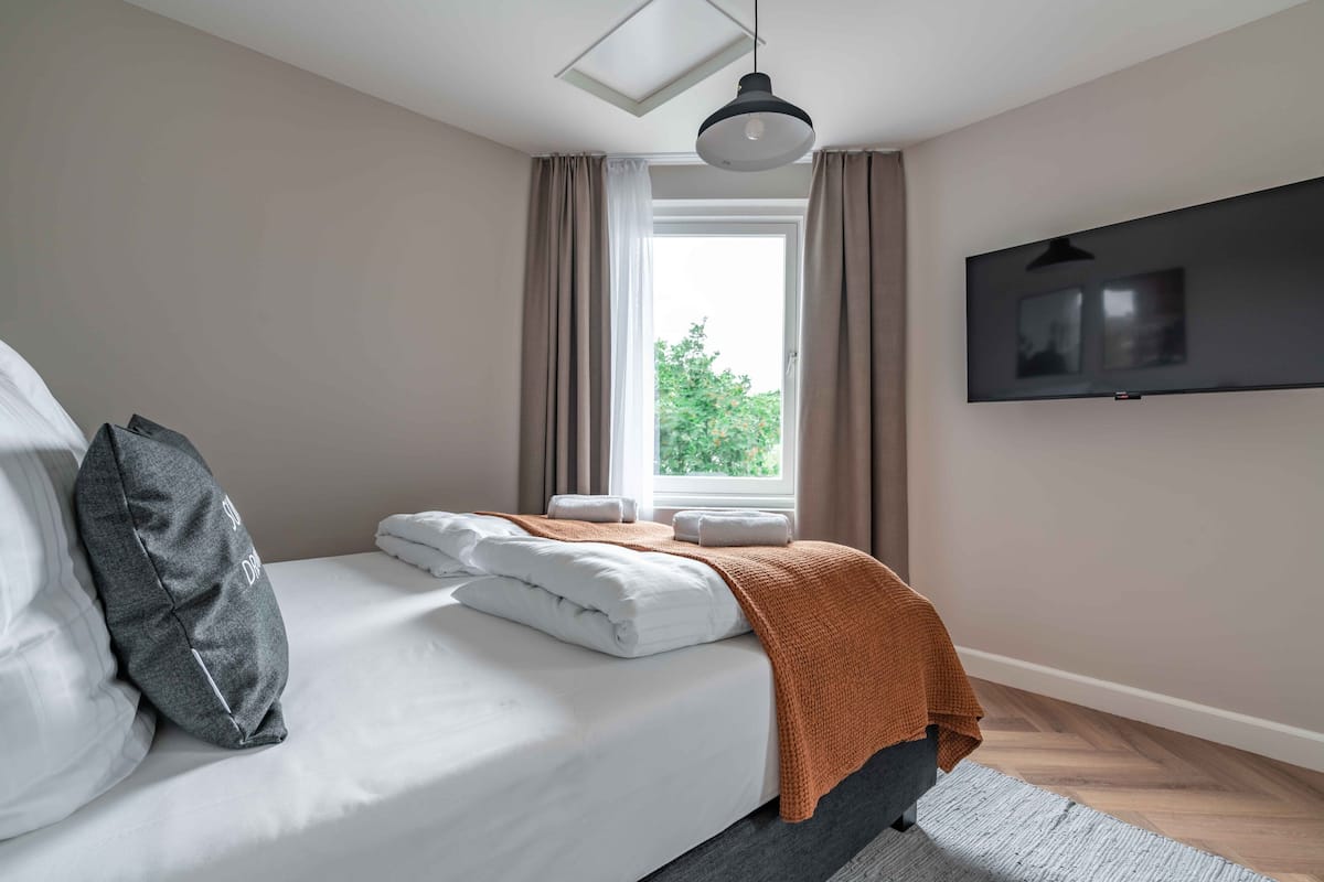 A neatly made bed with white linens and a brown throw blanket, a grey pillow, and a black pendant light above. A flat-screen TV is mounted on the wall to the right.