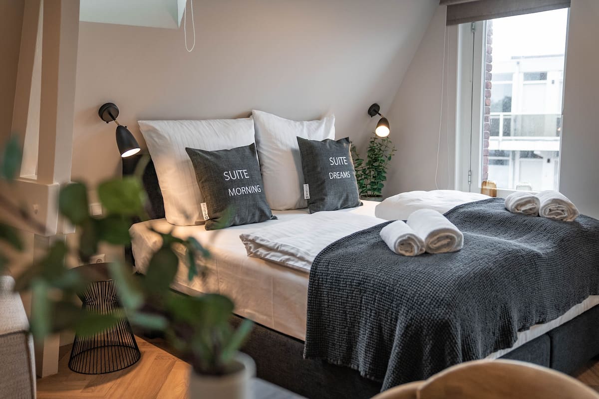 A modern bedroom with a bed, pillows, a bedside table, a lamp, and a window.