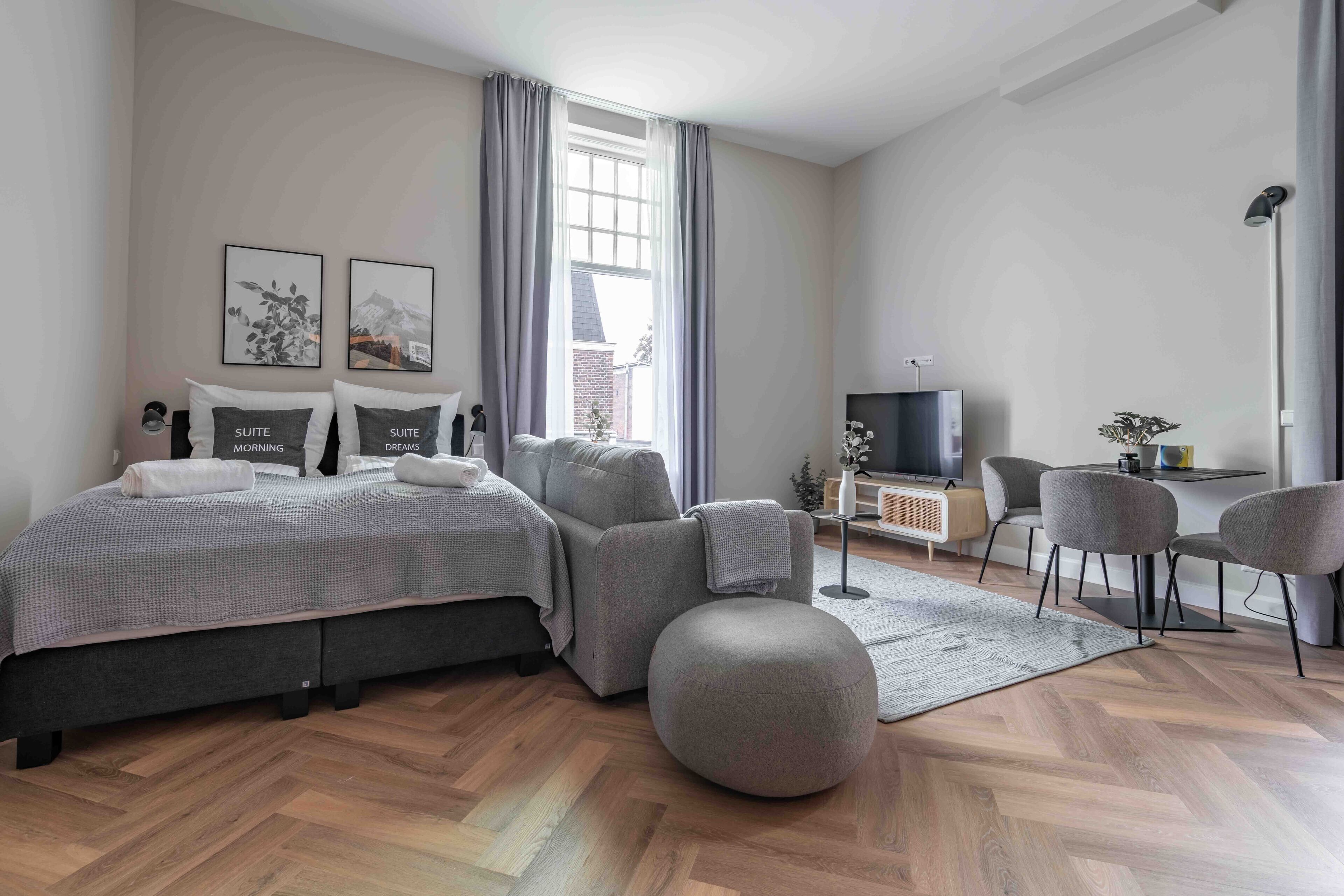 A modern bedroom with a large bed, grey upholstered chairs, a small desk, and a television.