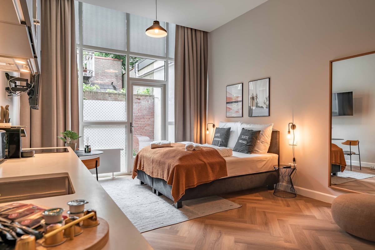 A modern bedroom with a large window, a bed with a brown blanket, a bedside table, and a mirror.
