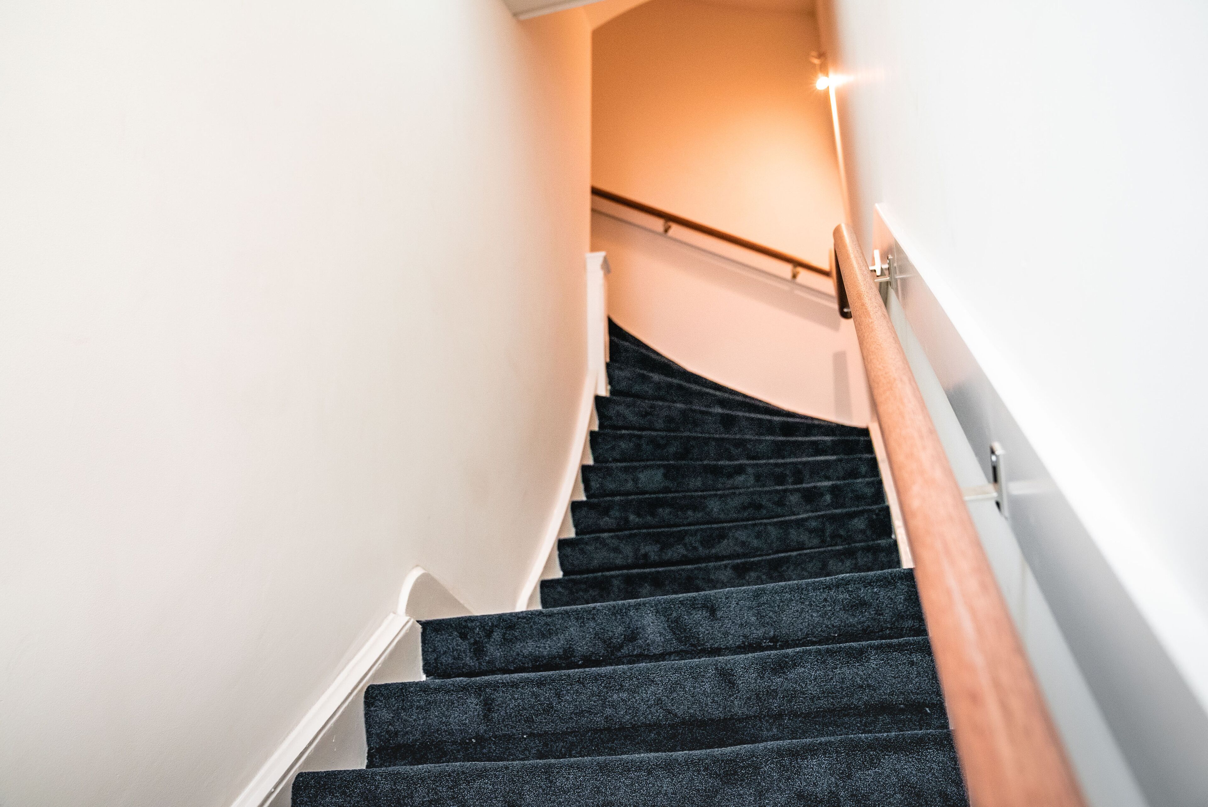 Un escalier avec des marches recouvertes d’une moquette sombre et une rampe en bois, menant à une pièce aux murs blancs et dotée d’un luminaire.