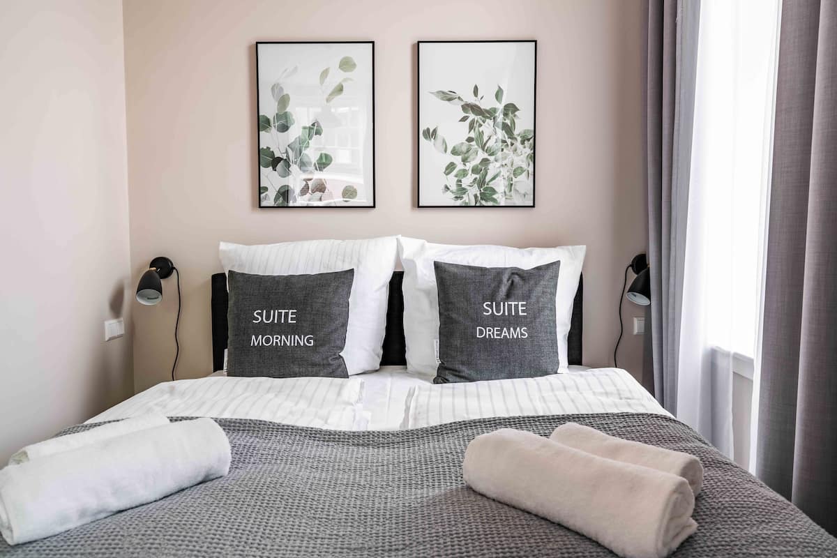 A neatly made bed with pillows labeled "SUITE MORNING" and "SUITE DREAMS," a window with curtains, and two wall-mounted lamps.