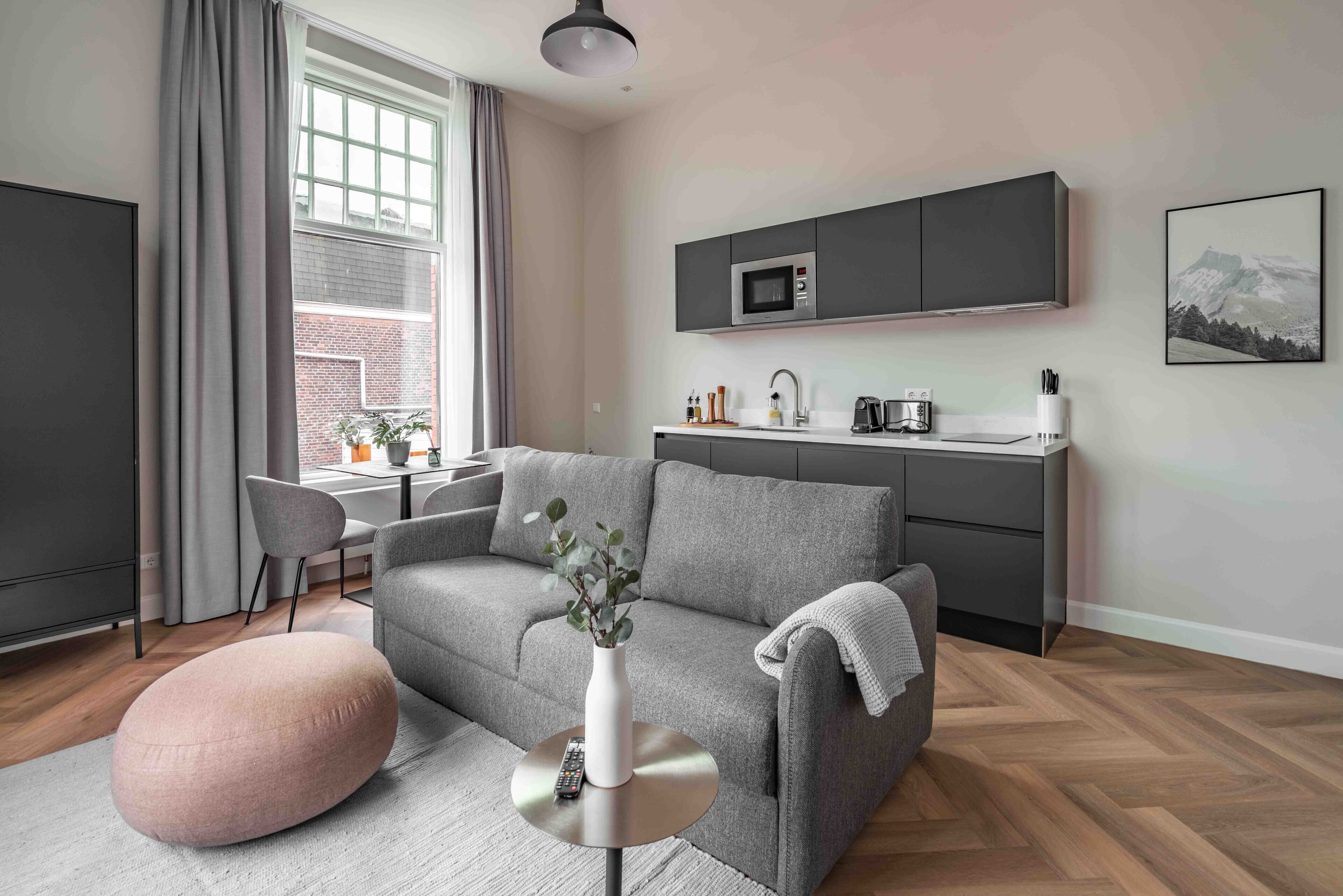 A modern living room with a grey sofa, a pink pouf, a white vase with a plant, and a kitchen area with a microwave and sink.