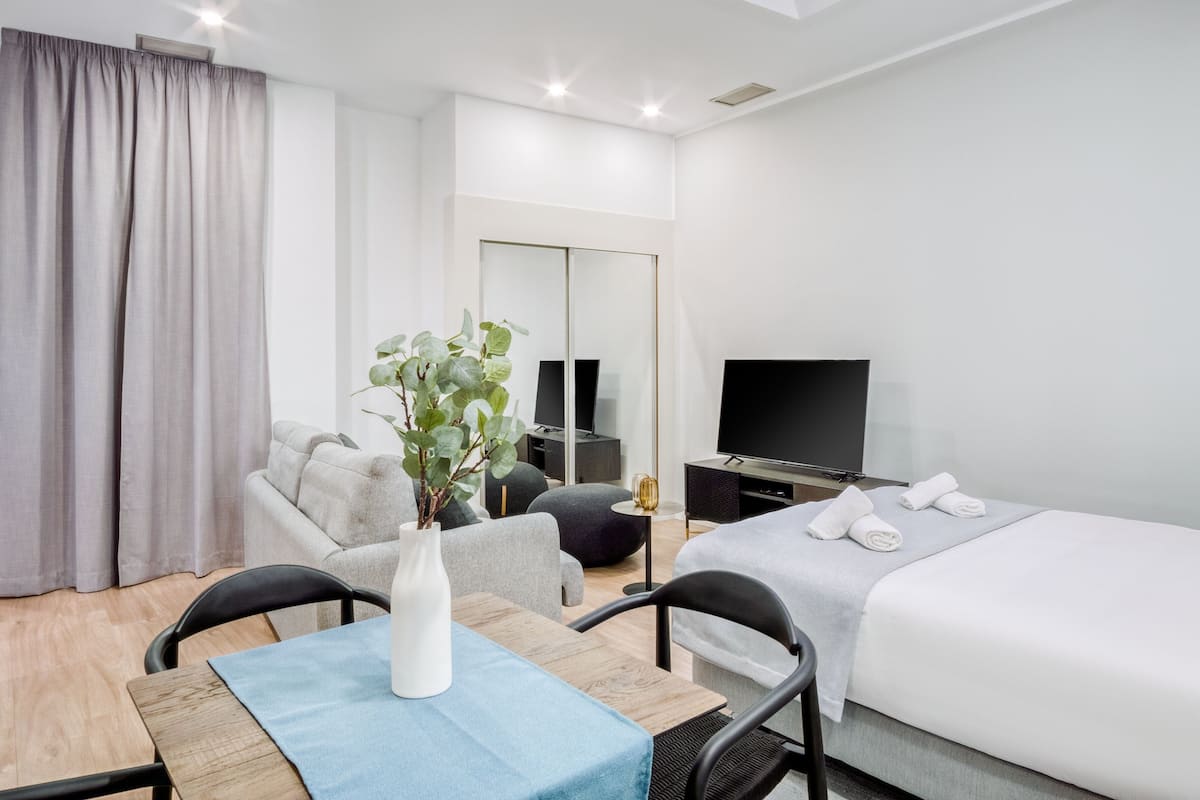A modern hotel room with a bed, a TV, a seating area with a table and chairs, and a large mirror.
