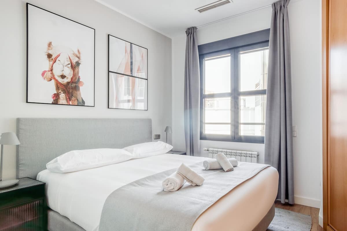 A modern hotel room with a large bed, a radiator, a window with curtains, and two framed artworks on the wall.