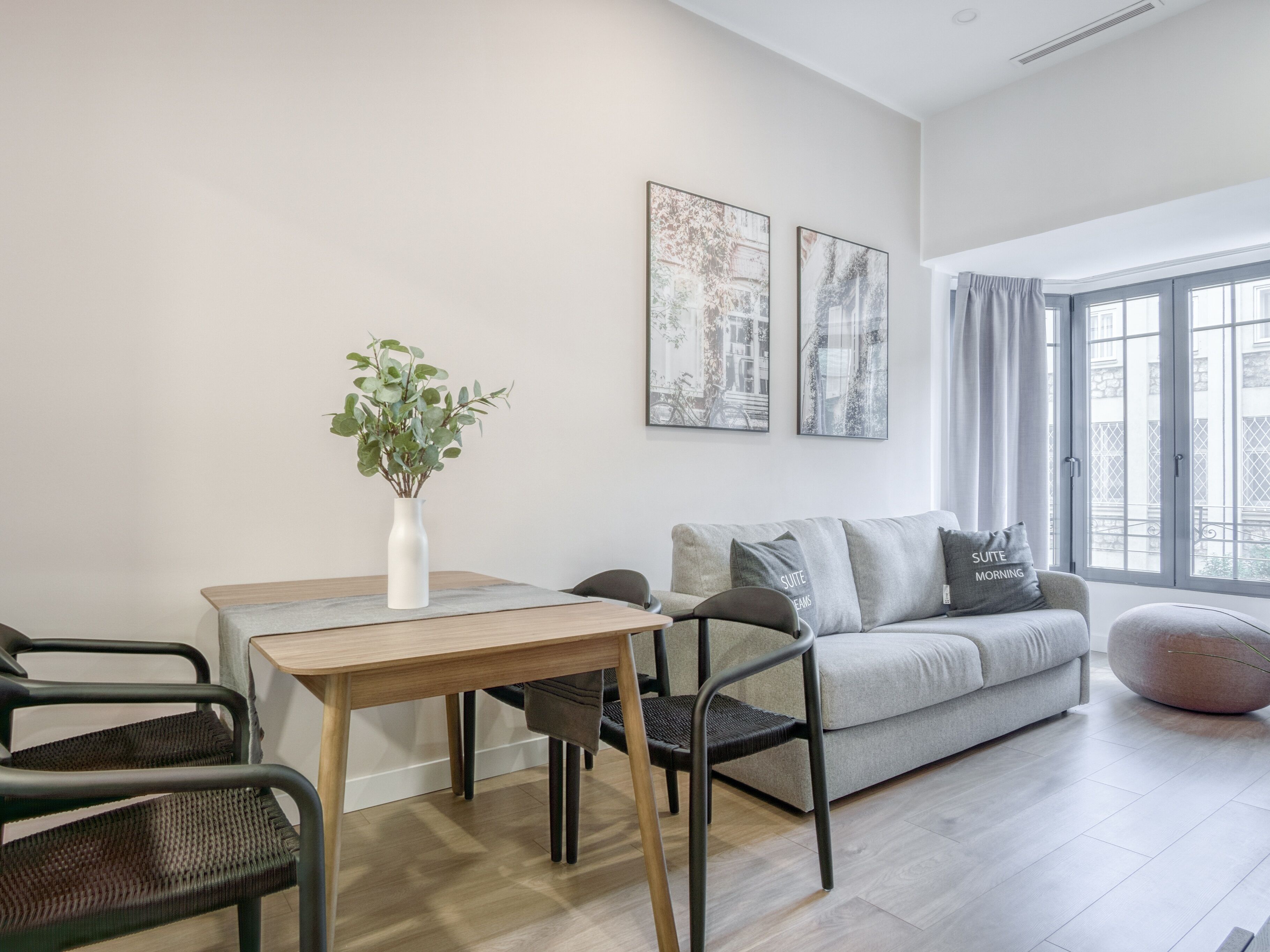 A modern living room with a grey sofa, a round ottoman, a dining table with a vase of green plants, and two framed pictures on the wall.