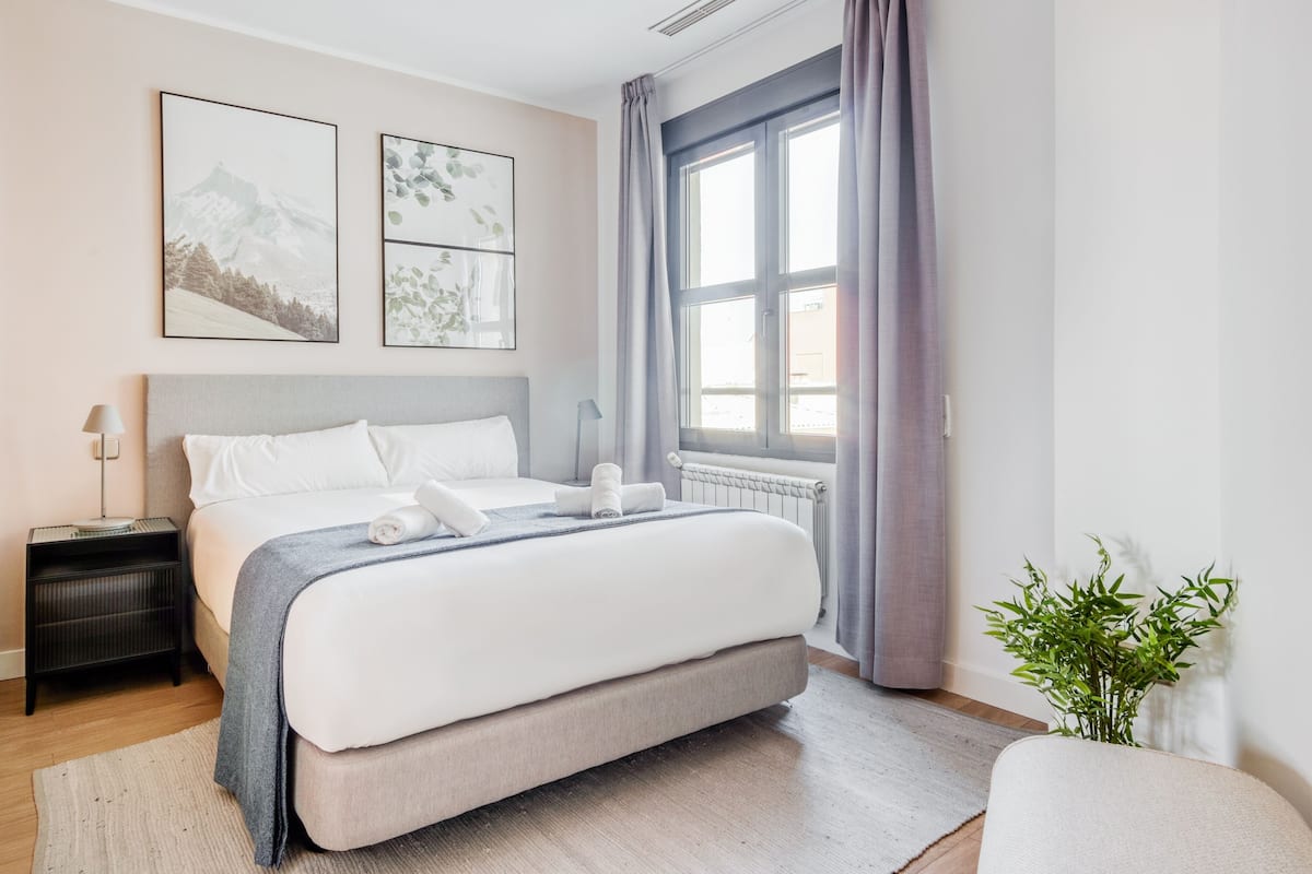 A modern bedroom with a bed, bedside tables, a window with curtains, and two framed pictures on the wall.