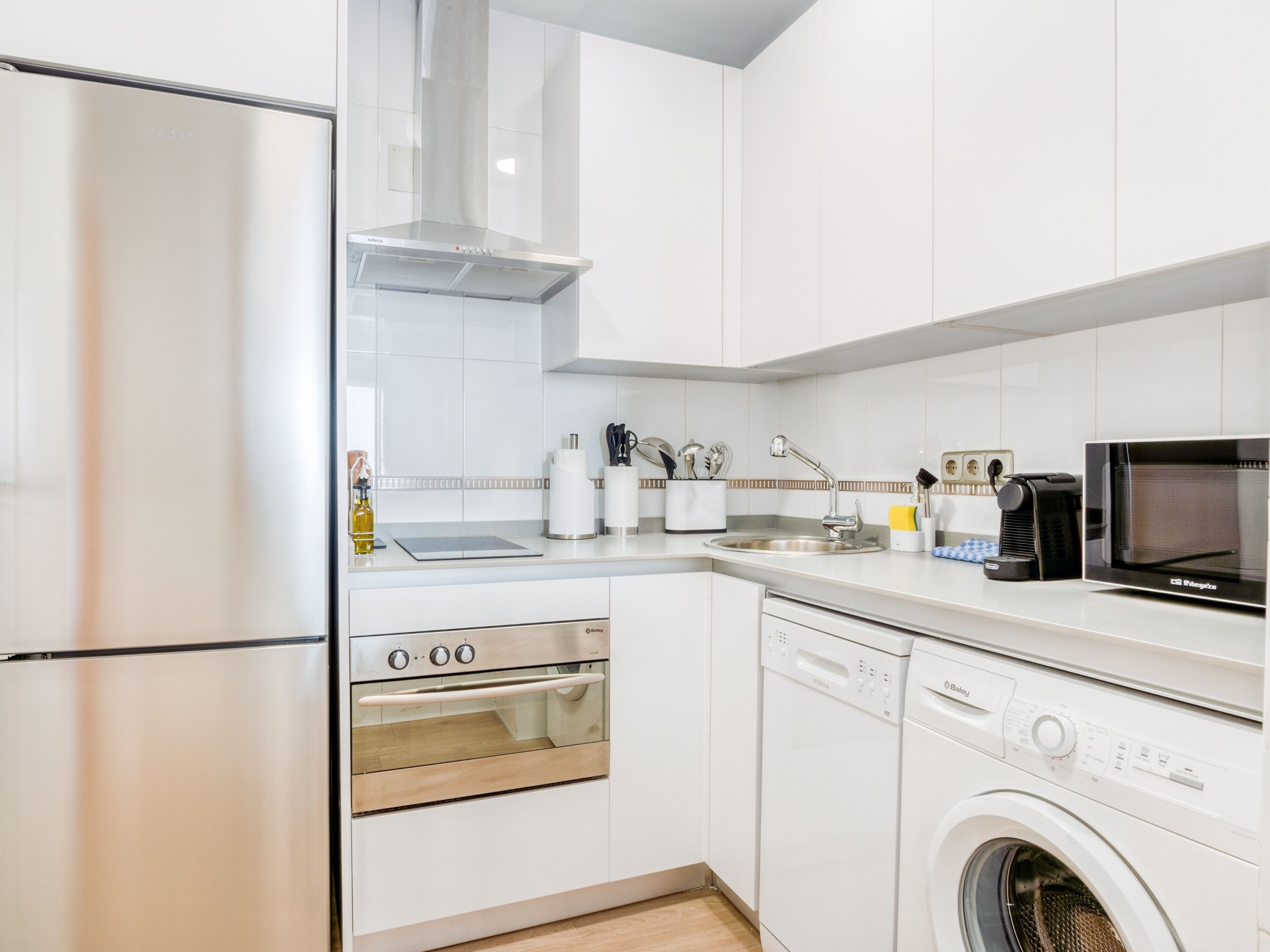 Een moderne keuken met witte kastjes en roestvrijstalen apparatuur, waaronder een koelkast, oven, vaatwasser en magnetron.