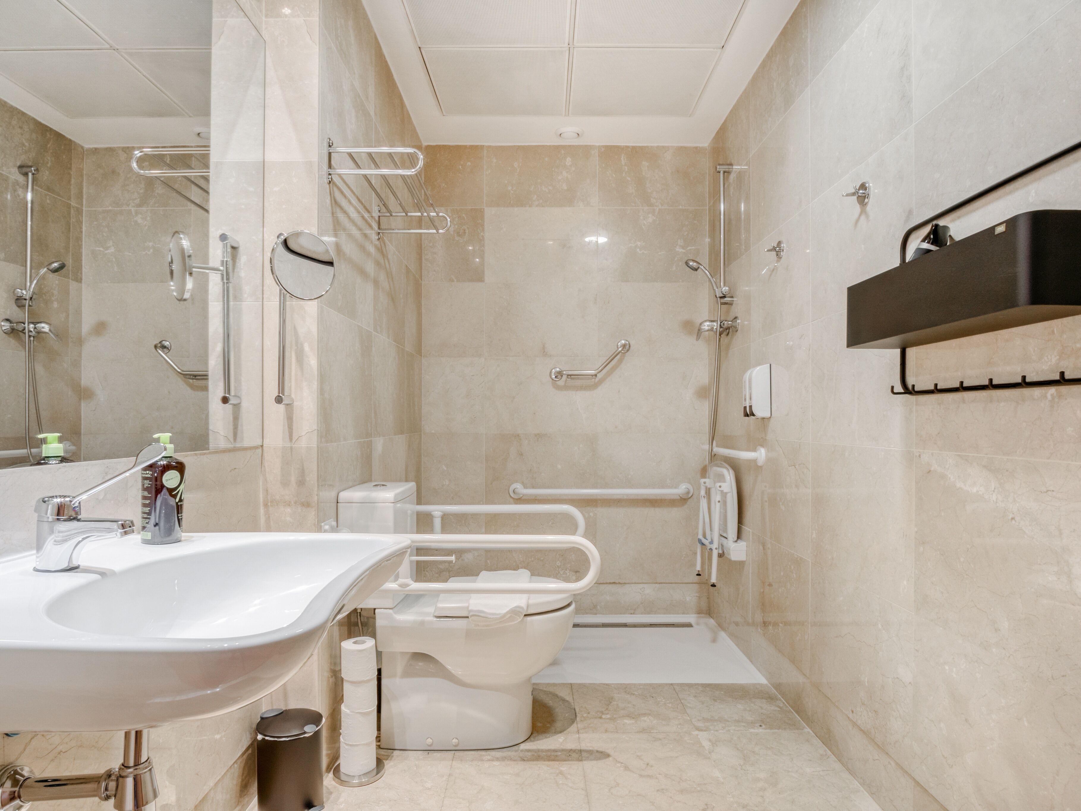 A modern bathroom with a sink, toilet, and shower area, including grab bars and a hand dryer.