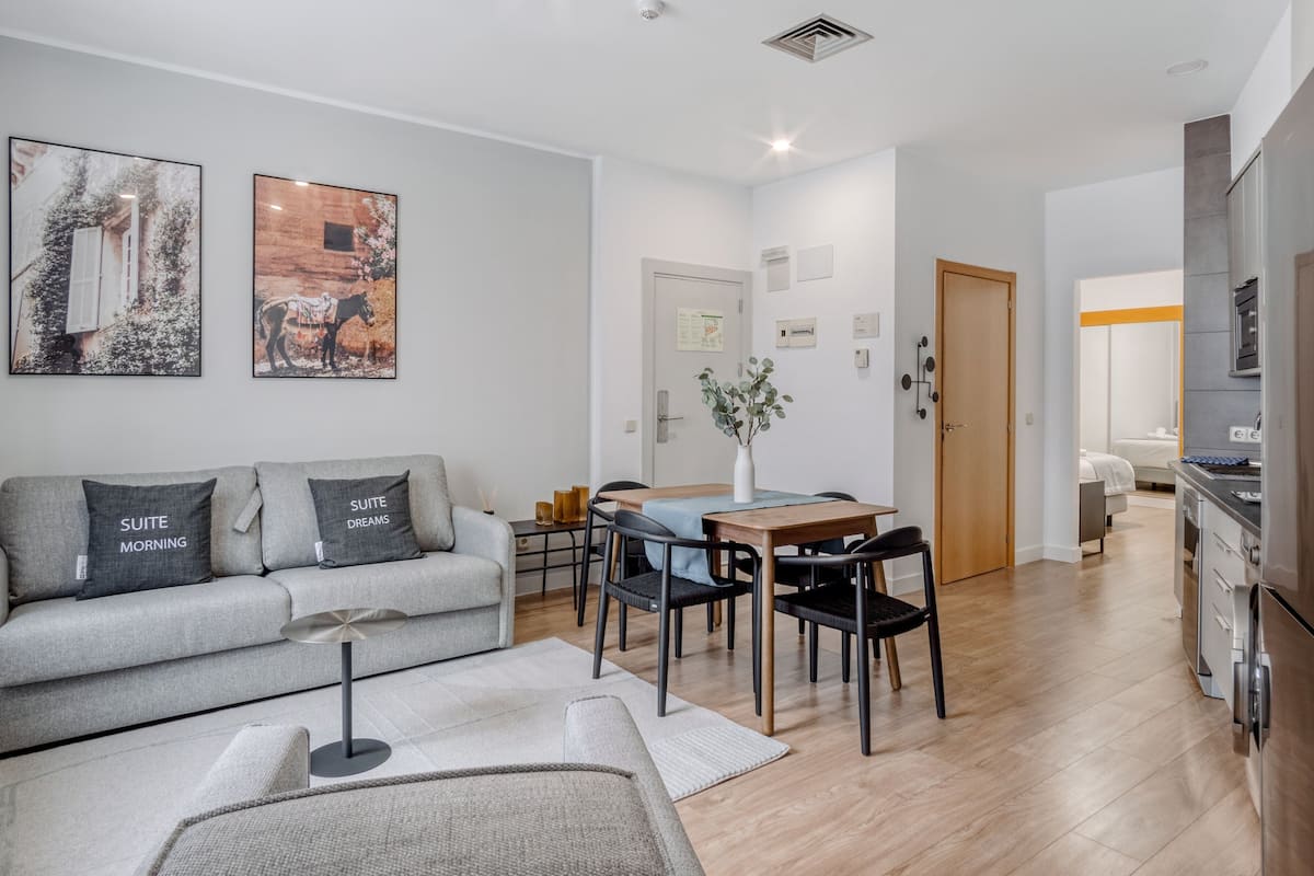 A modern living room with a grey sofa, a coffee table, and a dining area with a wooden table and chairs. There is a kitchen area with a refrigerator and a stove.