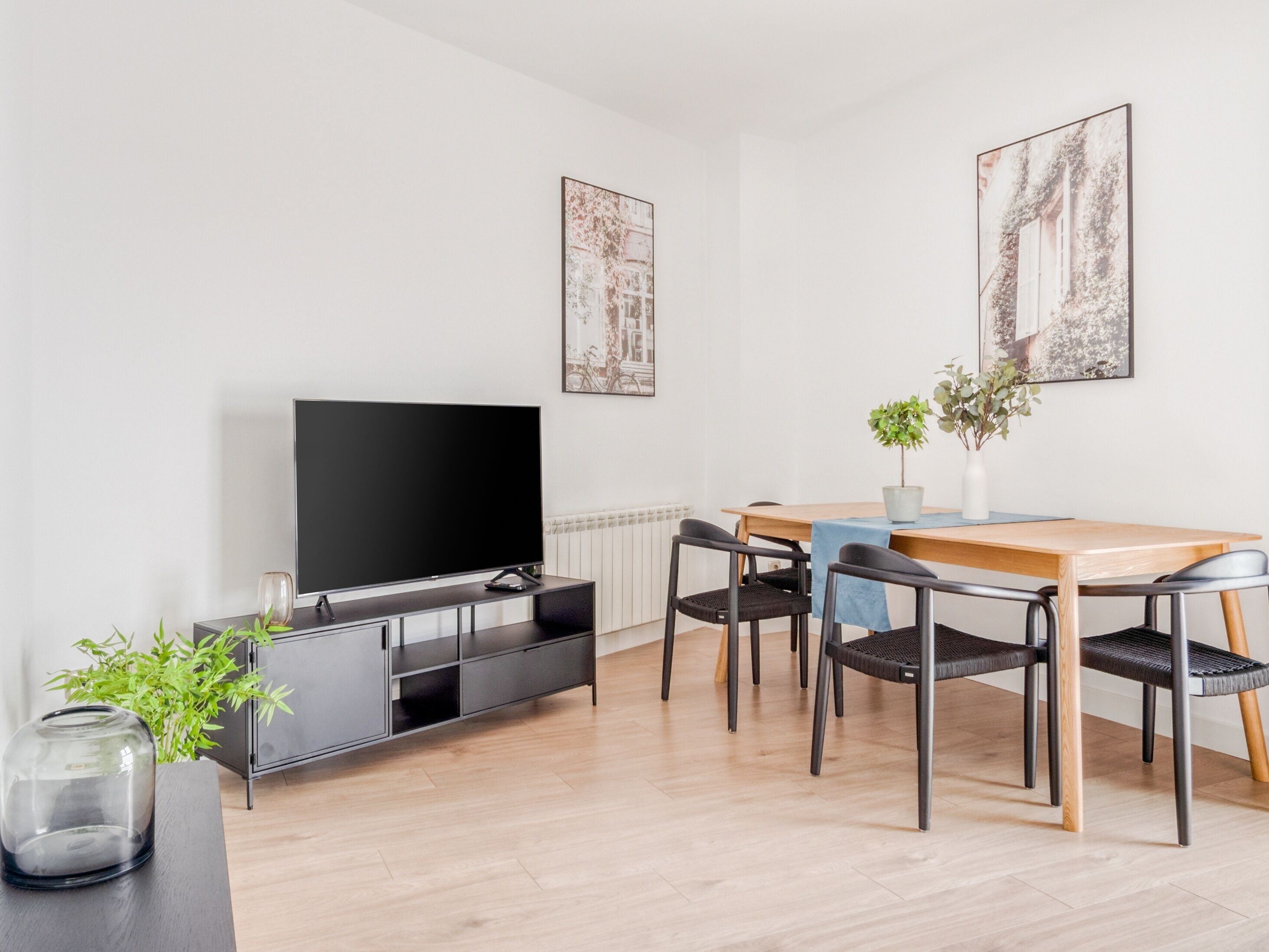 A modern living room with a flat-screen TV, a dining table with chairs, a potted plant, and two framed pictures on the wall.
