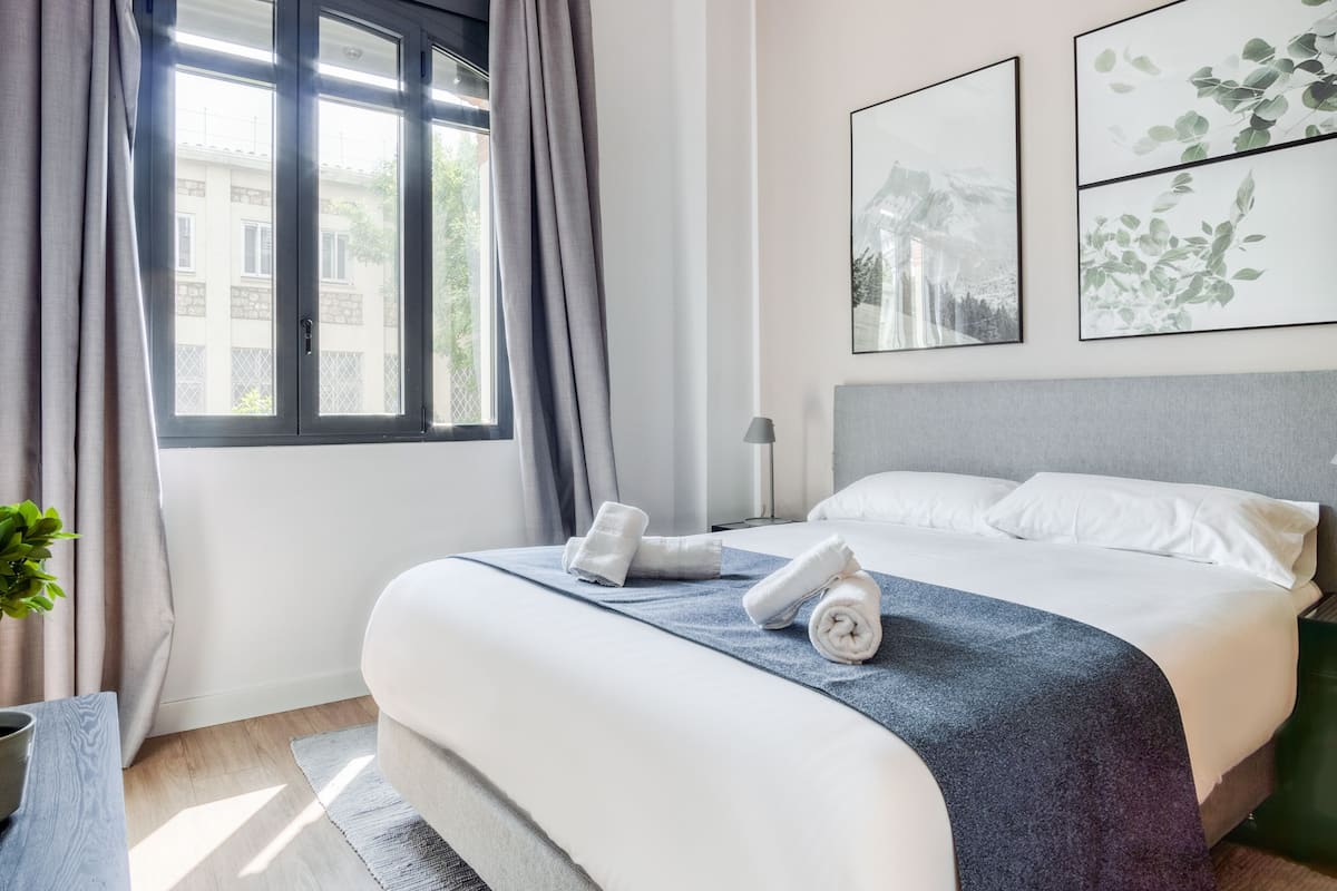 A modern hotel room with a large bed, a bedside table, a lamp, and a window with curtains.