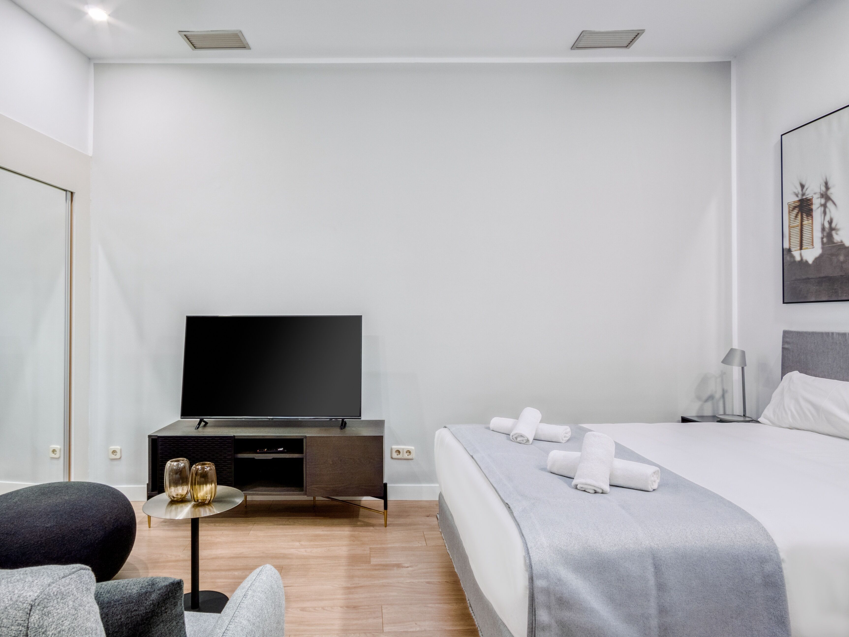 A modern hotel room with a flat-screen TV, a bed with neatly folded towels, a small side table with decorative vases, and a minimalist nightstand.