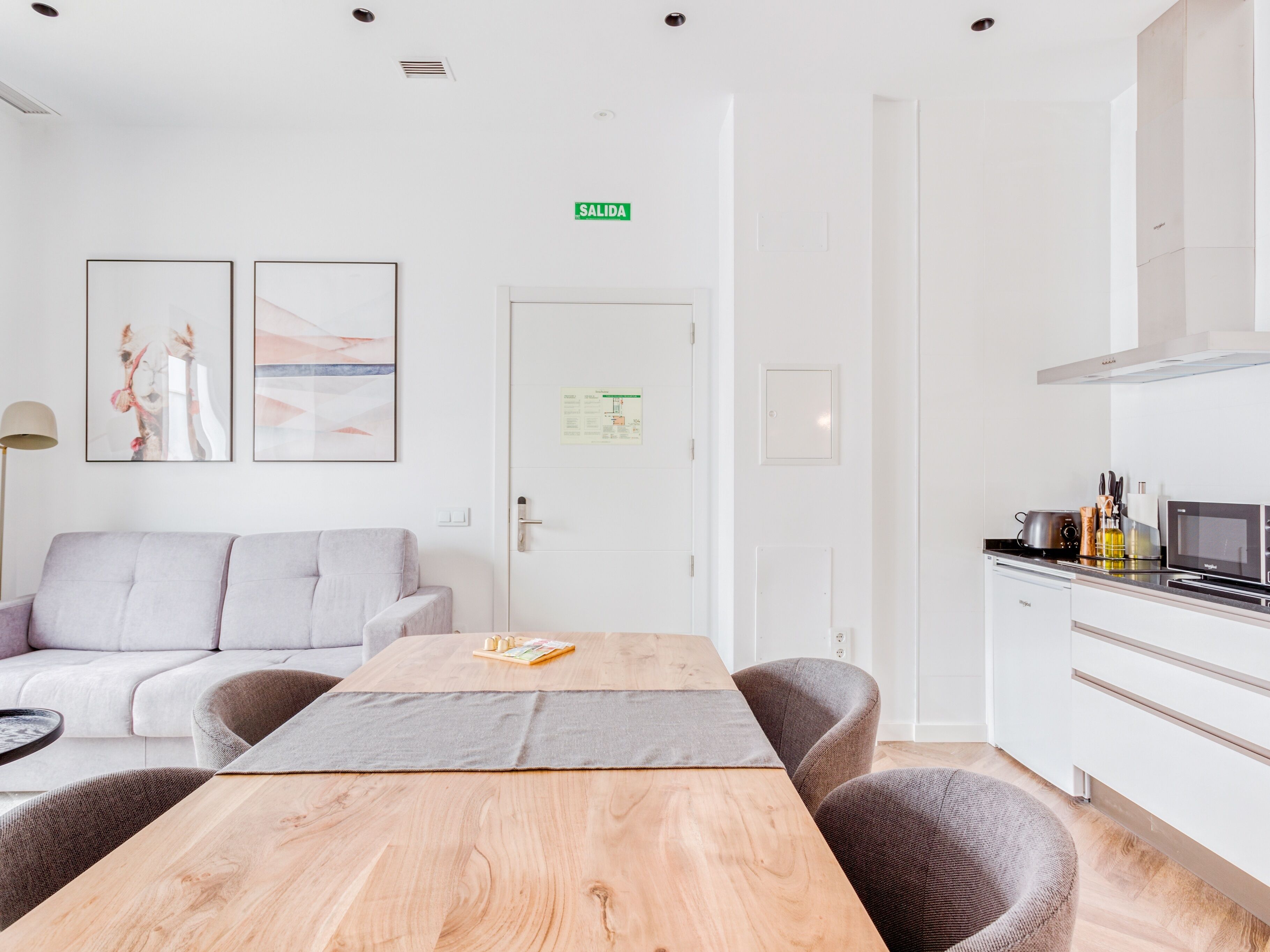 Ein moderner, minimalistischer Raum mit einem Holztisch, einem grauen Sofa und einer Küchenzeile inklusive Mikrowelle und Toaster.