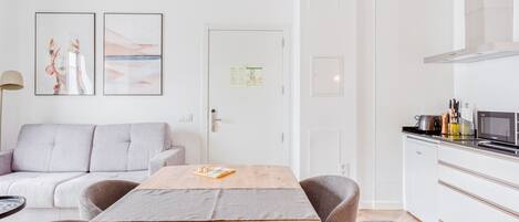 A modern, minimalist room with a wooden table, grey sofa, and a kitchenette with a microwave and toaster.