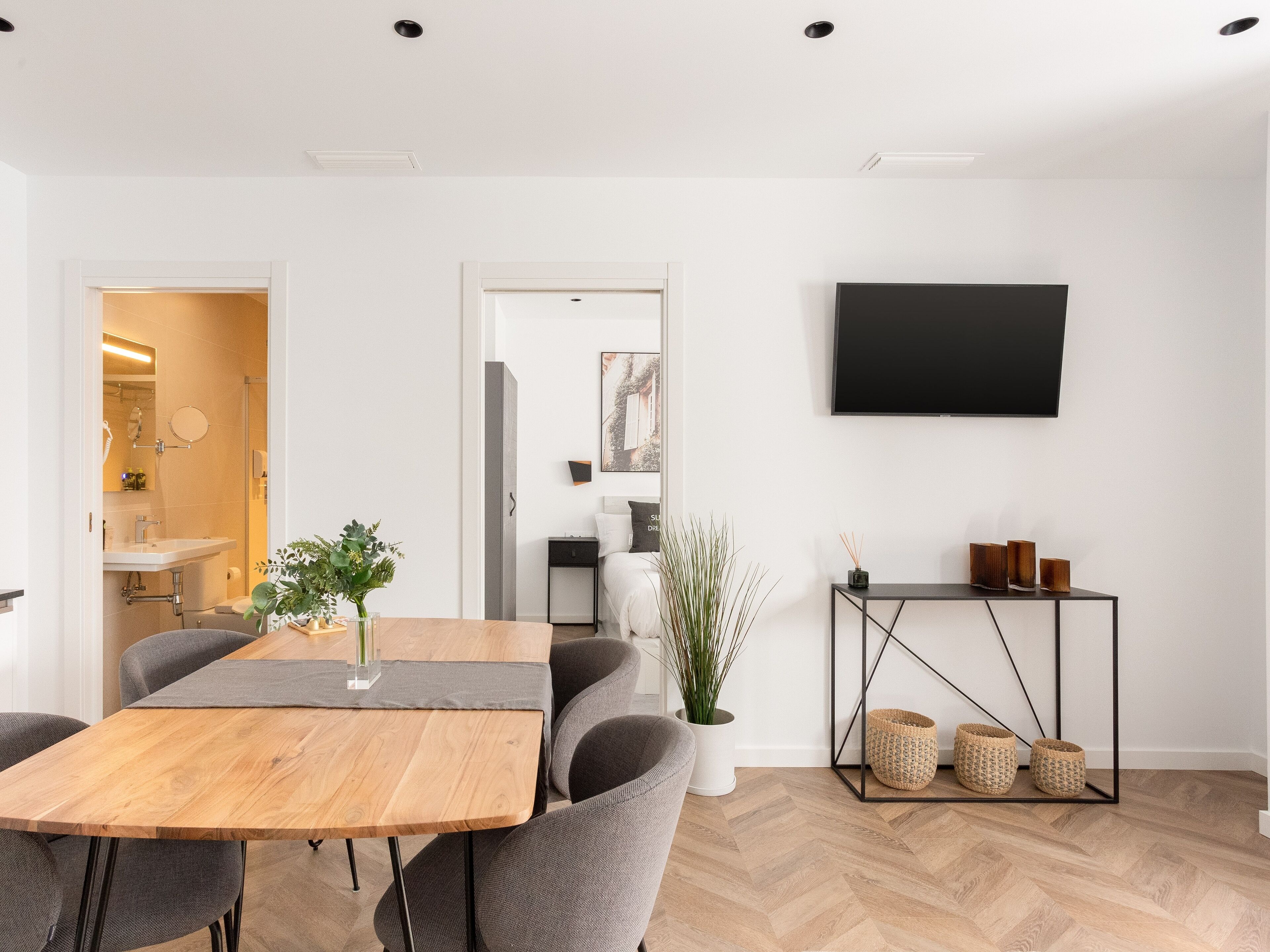 Een moderne woonkamer met een houten eettafel, grijs gestoffeerde stoelen, een flatscreen televisie aan de muur, een bijzettafeltje met decoratieve objecten en een potplant.