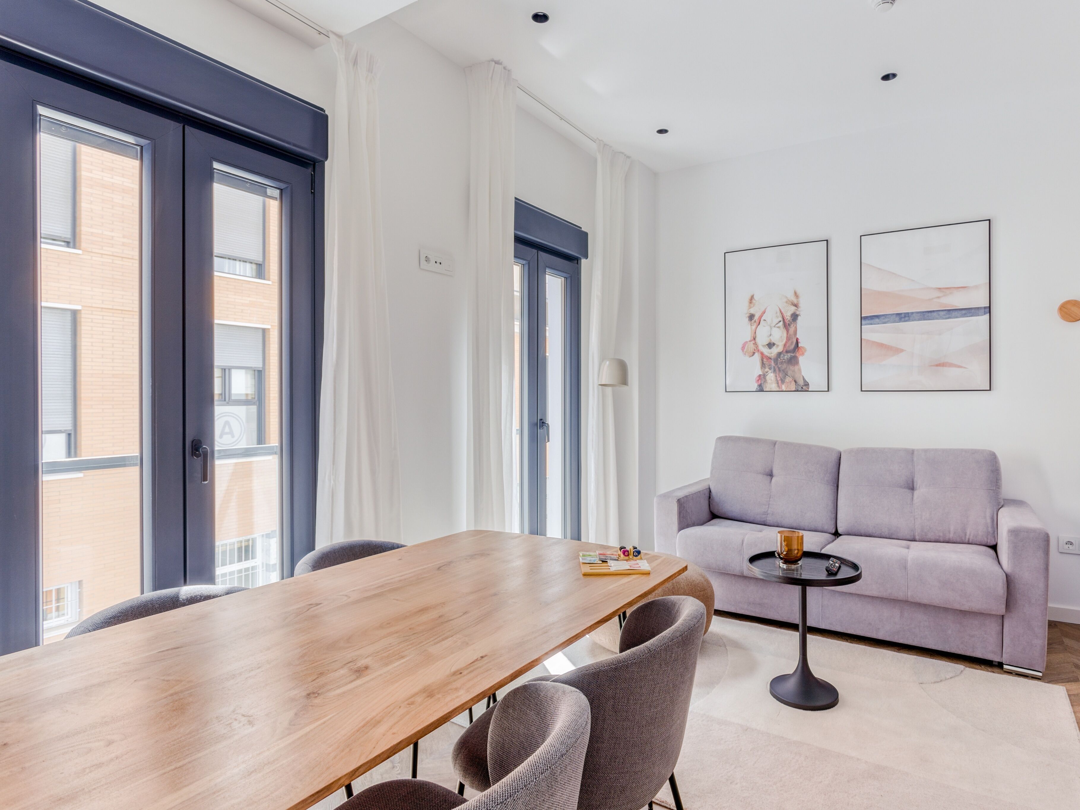 Un salon moderne avec une table à manger en bois, un canapé recouvert d’un tissu gris et deux œuvres d’art encadrées accrochées au mur.