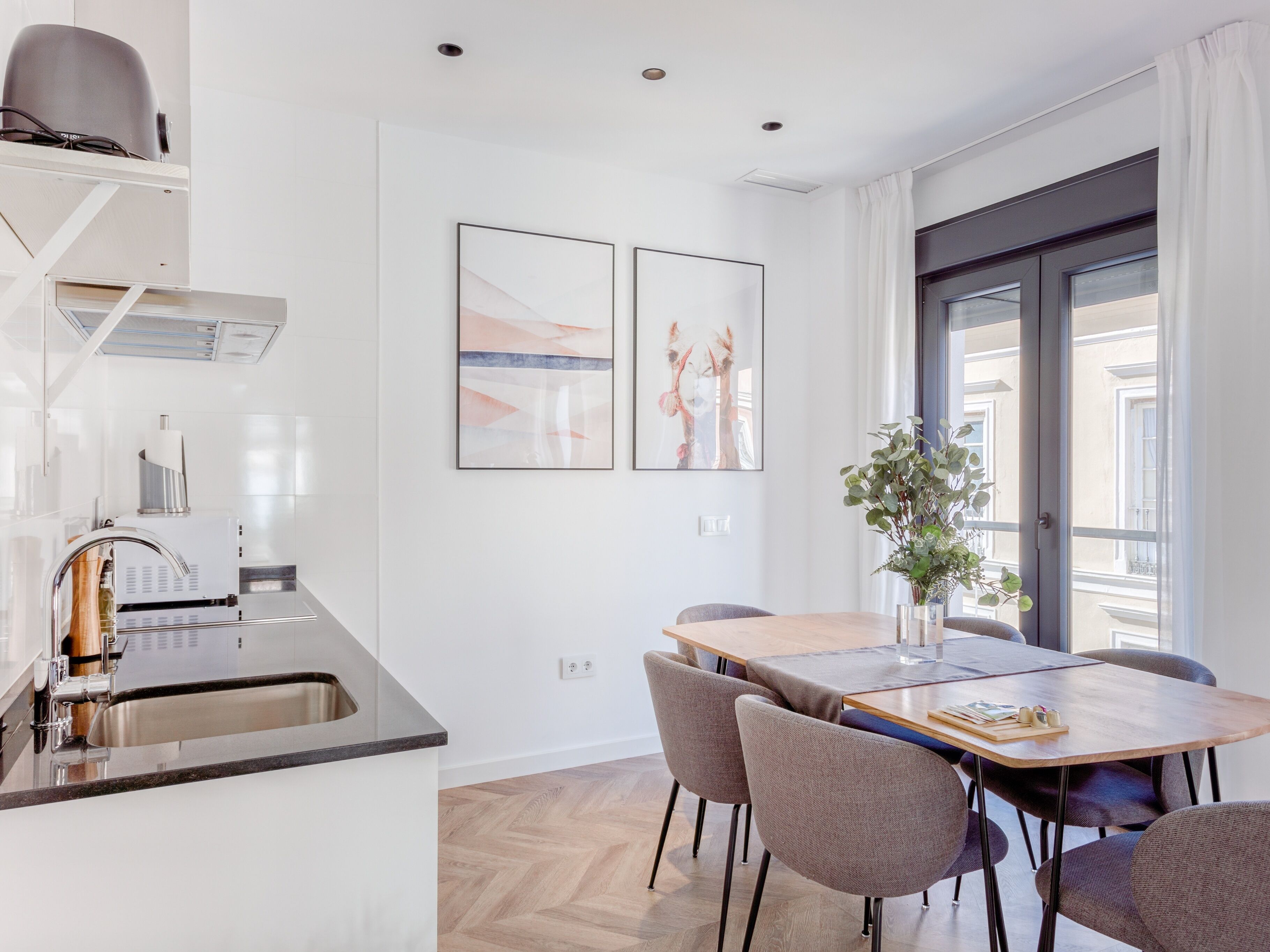 A modern kitchen with a built-in oven, a sink, and a stainless steel range hood. There is a dining area with a wooden table and six chairs. Two framed artworks are hung on the wall above the dining table.