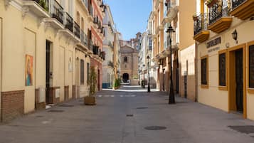 Una calle estrecha flanqueada por edificios, con balcones adornados con plantas y un cielo despejado.