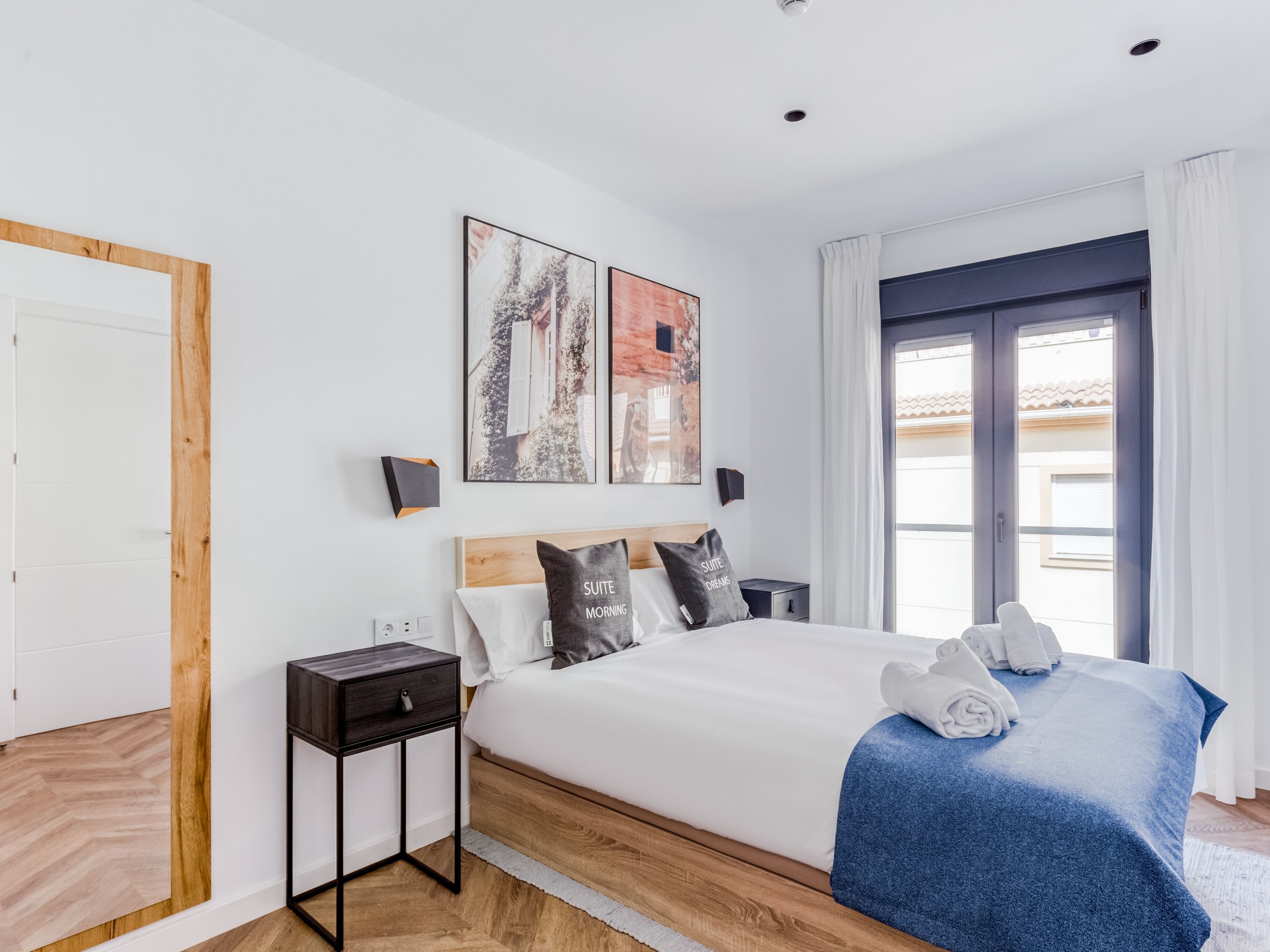 Een moderne hotelkamer met een groot bed, een houten hoofdeinde, twee kunstwerken aan de muur, een nachtkastje en uitzicht buiten door een raam.