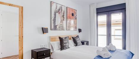 A modern hotel room with a large bed, a wooden headboard, two wall-mounted artworks, a nightstand, and a view of the outside through a window.