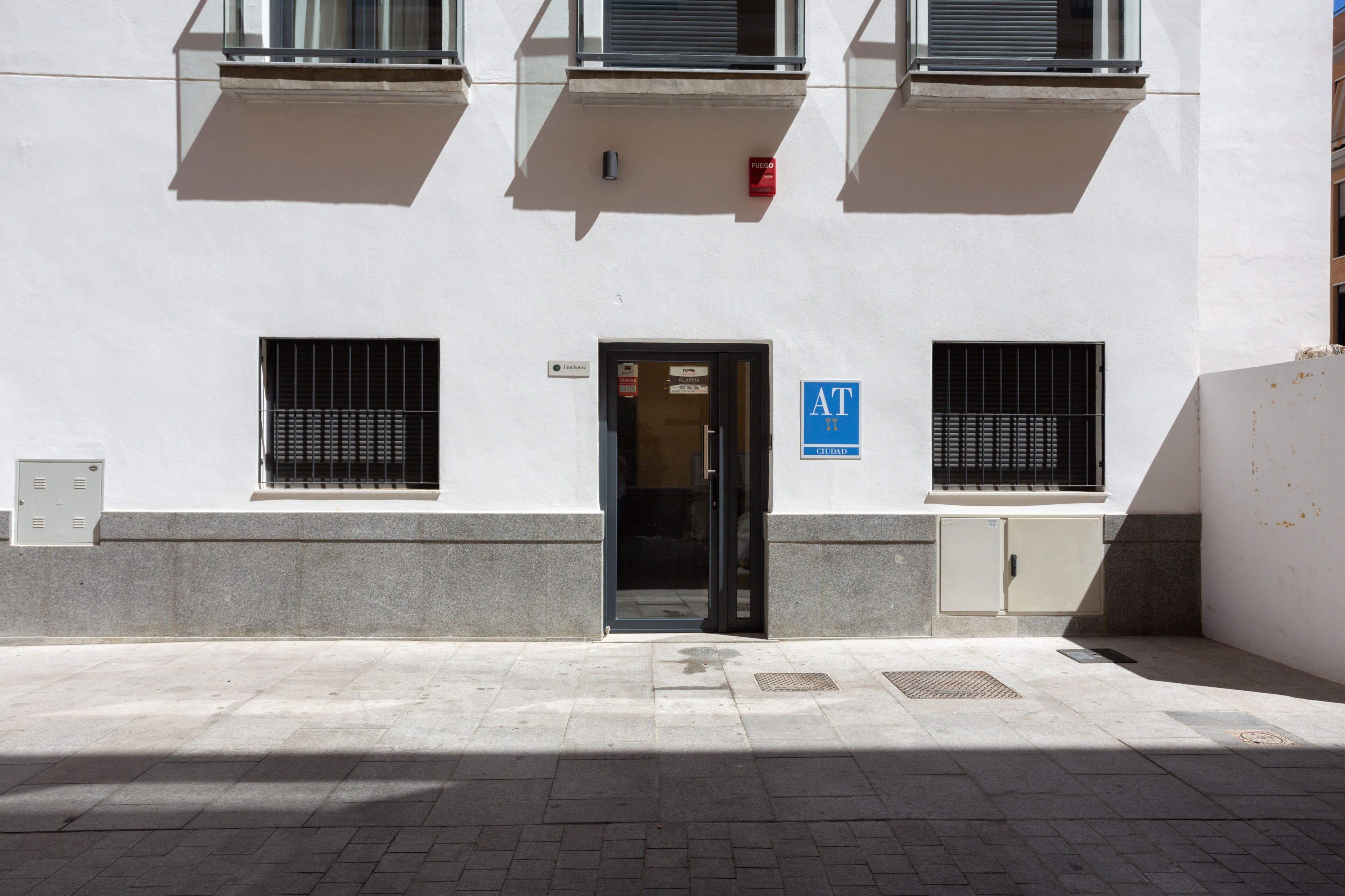 Fachada de edificio con una puerta negra, dos ventanas con rejas de metal, un letrero azul con los textos 'AT' y 'CITIDO', y una pequeña caja de servicios.
