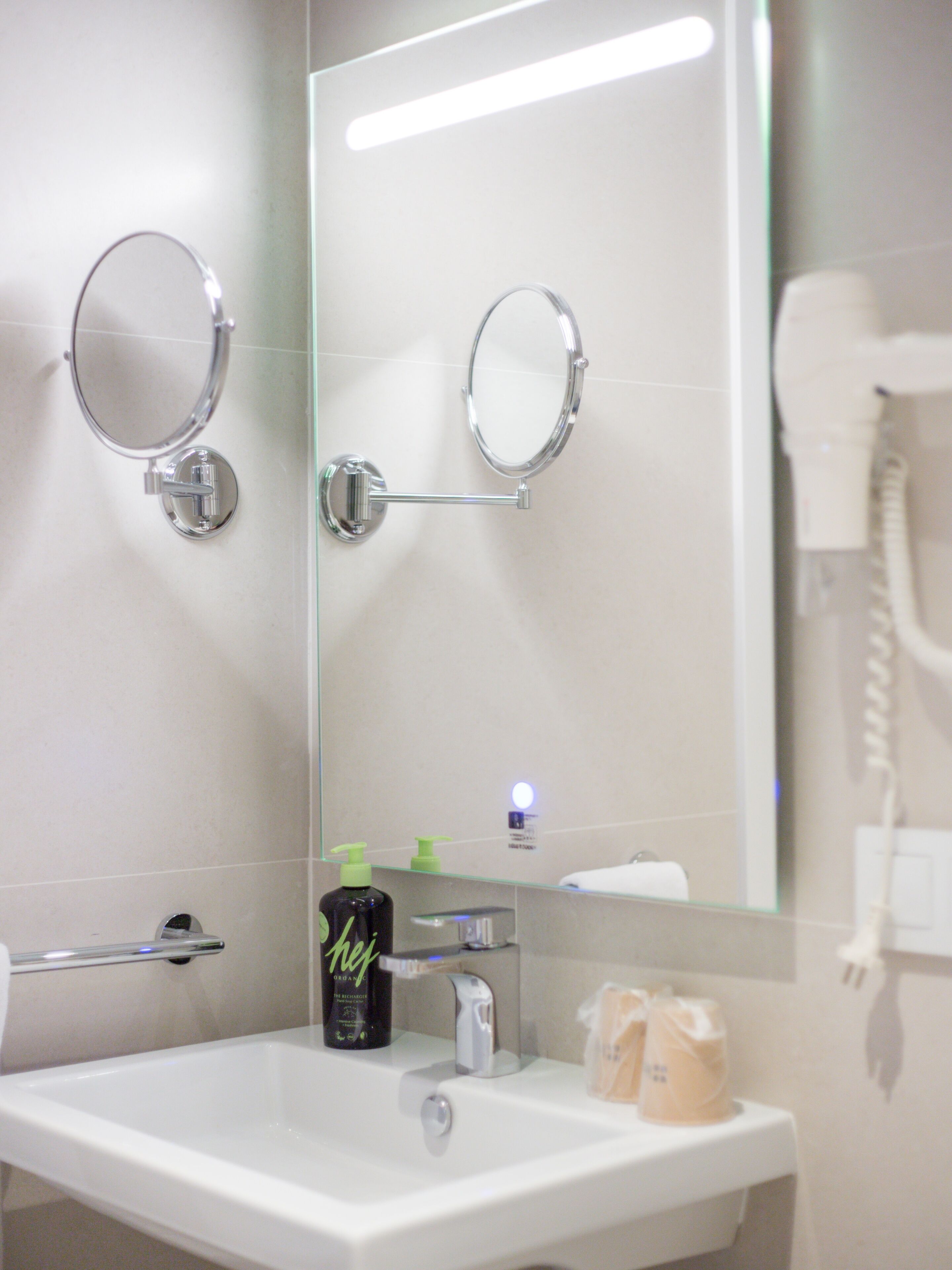 A bathroom with a white sink, two round mirrors, a hairdryer, and a bottle of shampoo.